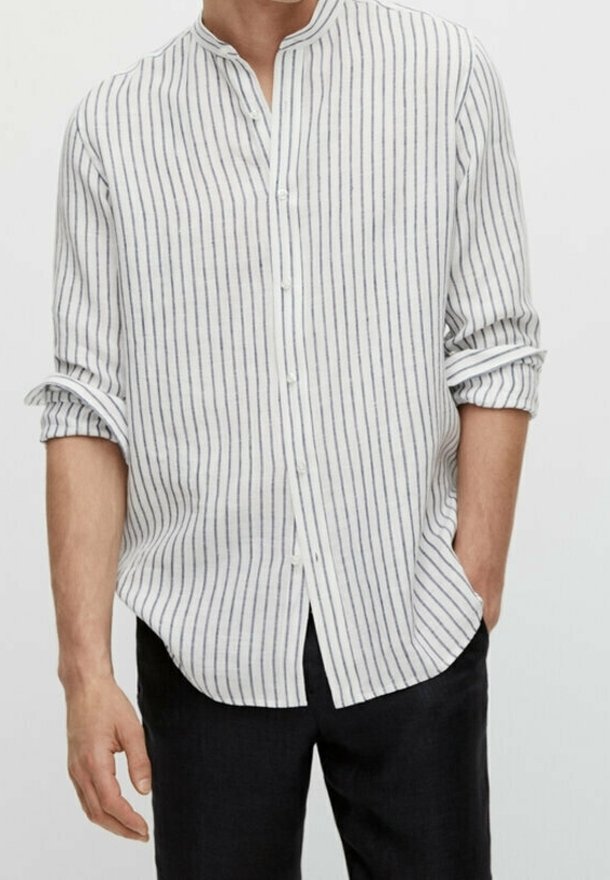 Witte linnen blouse met verticale marineblauwe strepen, mandarine kraag, lange mouwen opgerold en knopen aan de voorkant. Gedragen met donkere broeken.