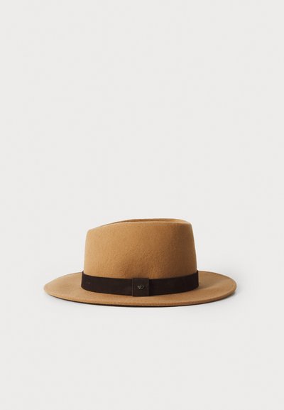 Chapeau marron en feutre avec un large bord et une bande en daim marron foncé. Présente un accent en métal carré sur la bande. Formé avec une couronne haute.