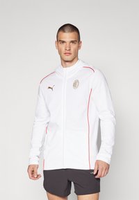 Hvid zip-up hoodie med røde detaljer og et guld logo på brystet, lavet af blødt stof, med lange ærmer og hætte.