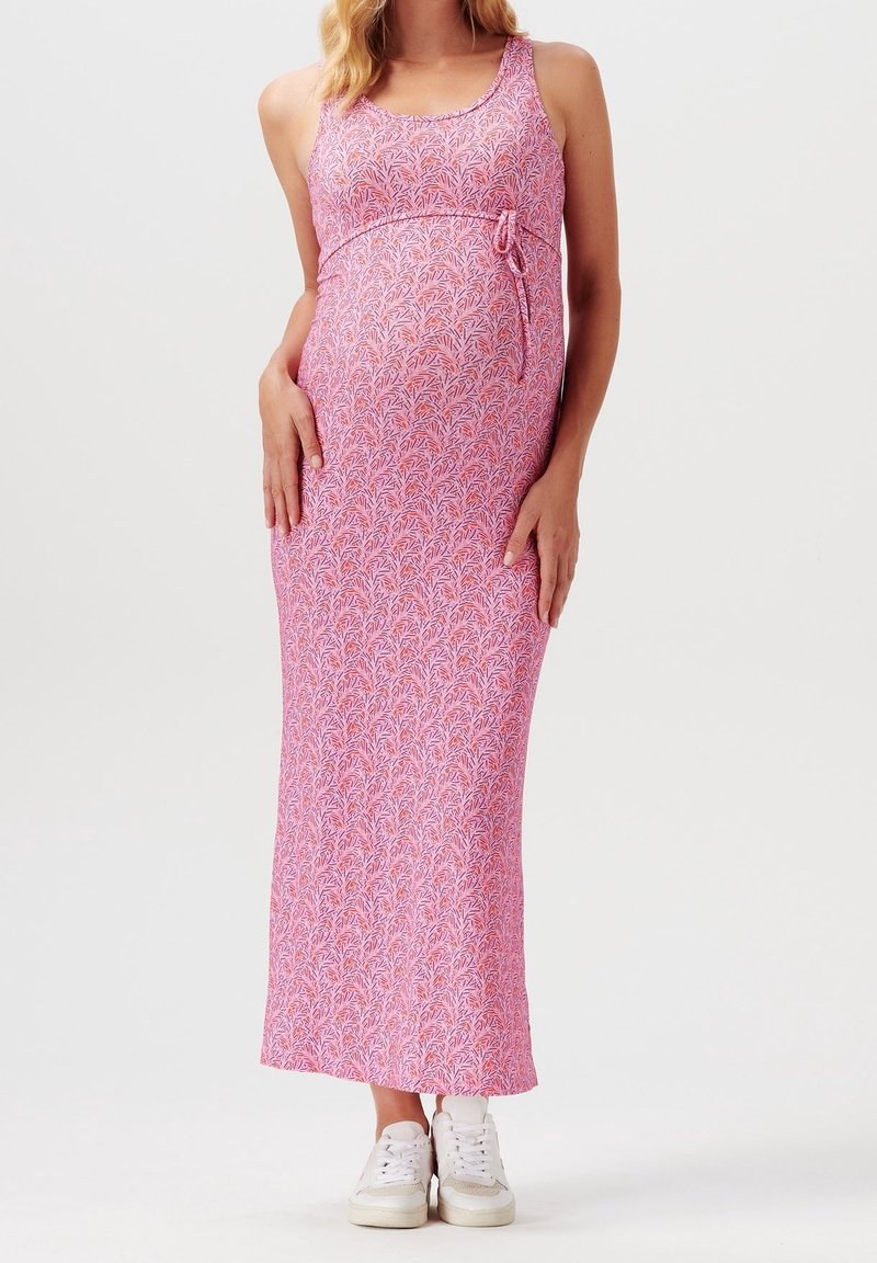 Robe maxi de maternité rose avec un motif de feuilles, sans manches, lien ajustable à la taille, en tissu doux, associée à des baskets blanches.