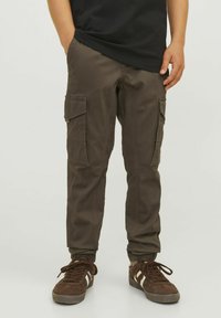 Olive green cargo nohavice z bavlneného zmesi, ktoré majú viacero bočných vreciek, zúžené nohavice a elastické manžety. Skombinované s hnedými teniskami.