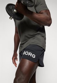 Calções atléticos pretos com a inscrição "BORG" em letras brancas, combinados com uma t-shirt justa verde-azeitona, segurando um haltere na mão.