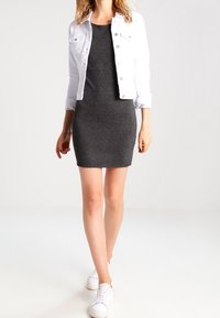 Vestito grigio aderente in materiale elasticizzato, abbinato a un giubbotto di jeans bianco. Modella con sneakers bianche. Sfondo semplice.