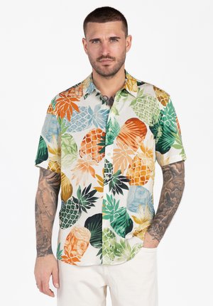 Mężczyzna o krótkich włosach i tatuażach, ubrany w kolorową, krótką koszulę na guziki z dużymi wzorami ananasów i liści.