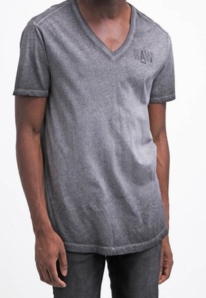 Homme portant un T-shirt gris délavé en col V avec l'inscription "RAW" imprimée sur la poitrine gauche, associé à un pantalon gris foncé.