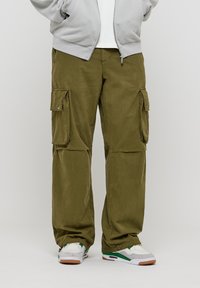 Pantalon cargo en coton vert olive, avec poches latérales, coupe ample et texture subtile. Associé à des baskets blanches et vertes.