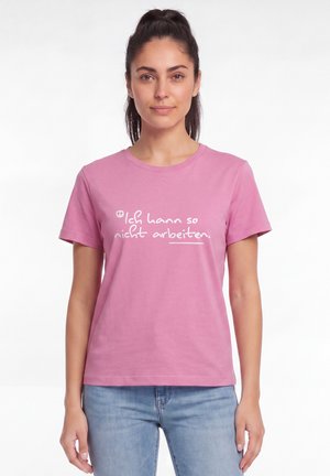 Junge Frau mit dunklen Haaren im Pferdeschwanz, die ein pinkes T-Shirt mit weißem deutschem Text und hellblaue Jeans trägt, steht vor einem weißen Hintergrund.