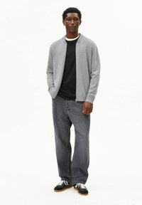 Graue Bomberjacke mit Reißverschluss über einem schwarzen T-Shirt, kombiniert mit lockeren dunkelgrauen Jeans und schwarzen Schuhen mit weißen Schnürsenkeln. Schlichter Hintergrund.