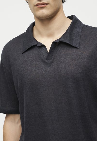 Άνδρας που φοράει σκούρο γκρι πόλο με κοντά μανίκια και ανοιχτό V-λαιμόκοψη, εμφανίζεται από το στήθος μέχρι το λαιμό.