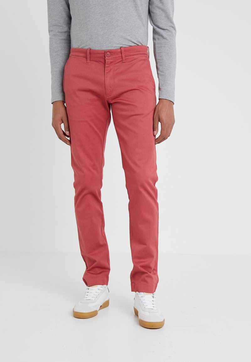 Homme portant un pantalon slim couleur corail, une chemise grise à manches longues et des baskets blanches avec semelles en gomme, debout sur un sol blanc.