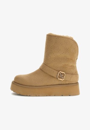 Stivale alla caviglia in suede tan con motivo embossé, fodera in pelliccia, cinturino largo con accento in metallo color oro a forma esagonale, e suola spessa.