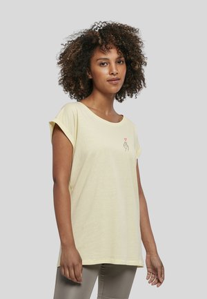 Femme aux cheveux bouclés portant un t-shirt jaune pâle avec un petit motif main et cœur sur la poitrine, debout devant un fond uni.