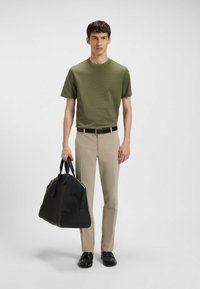 Groen gestructureerd t-shirt, beige broek, zwarte riem, houdt een grote zwarte tas met gestructureerde handvatten vast. Schoenen: zwart, slip-on ontwerp.