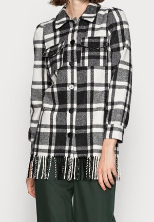 Czarno-biała koszula w kratę na guziki z kieszeniami na piersi i frędzlami na dole, noszona na ciemnozielonych spodniach, skrócona przy szyi i udach.
