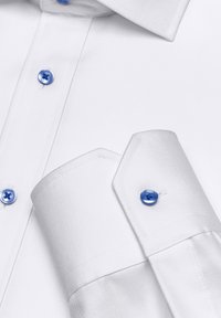 Chemise blanche en coton avec une texture lisse, présentant des boutons bleus au niveau du col et des poignets. Les poignets ont un design pointu.