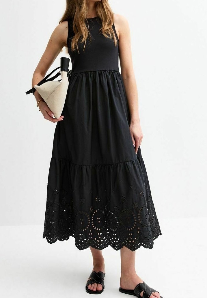 Femme portant une robe noire sans manches avec des détails œillets à l'ourlet, tenant un sac à main beige et noir, et portant des sandales noires à enfiler.