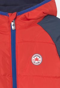 Czerwona kurtka puchowa z niebieskim zamkiem błyskawicznym i granatowymi akcentami. Z przodu znajduje się okrągła naszywka z logo Converse. Gładka, błyszcząca faktura.