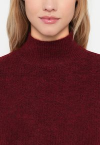 Bordo pulover s turtleneck o vratu, z rebrasto teksturo, izdelan iz mehkega materiala, s prilegajočim se dizajnom in visokim ovratnikom.