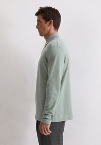 Polo verde deschis, cu mânecă lungă și guler. Fabricat dintr-un material moale, având o croială dreaptă și un design minimalist.