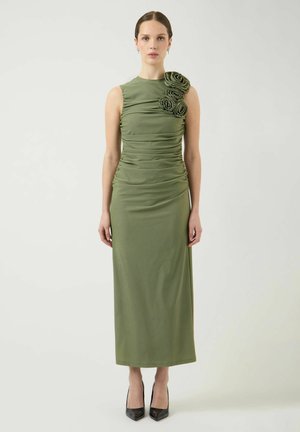 Naine õlgu paljastava oliivrohelise midi-kleidi ja kolme roosikujulise detailiga ühel õlal, seisab sirgelt, kandmas musti kontsakingi.