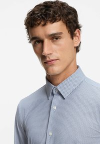 Blauw-wit geruit overhemd met knopen en een puntige kraag, voorzien van een gestructureerde stof en een subtiel patroon. Witte knopen zijn zichtbaar.