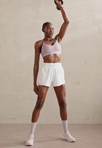 Sujetador deportivo rosa con logo blanco, pantalones cortos deportivos blancos, zapatillas rosas, levantando una kettlebell negra contra un fondo neutro.