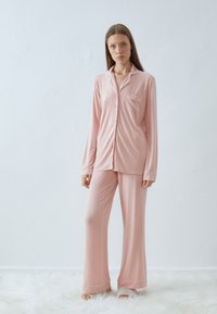 Ljust rosa satinpyjamas-set med lång ärm, krage, bröstficka och vida byxor med fåll-accenter. Mjuk textur.