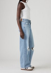 Světle modré džínsy s vysokým pasem a širokými nohavicemi, s opotřebovanými detaily na kolenou a klasickým střihem. Kombinovány s přiléhavým bílým tílkem a ležérní obuví.