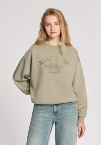 Khaki sweatshirt z okrągłym dekoltem, opuszczonymi ramionami i haftowanym napisem. Połączony z jasnoniebieskimi dżinsami. Miękka tekstura i luźny krój.