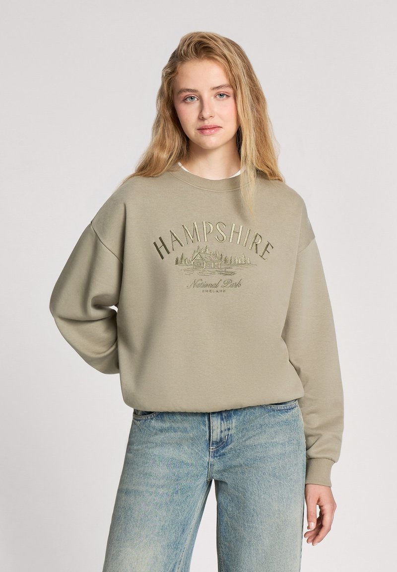 Khaki sweatshirt z okrągłym dekoltem, opuszczonymi ramionami i haftowanym napisem. Połączony z jasnoniebieskimi dżinsami. Miękka tekstura i luźny krój.