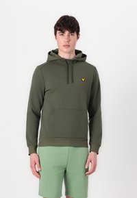 Maslinasto zelena hoodie s prednjim džepom, podesivom kapuljačom, rebrastim manžetama i malim žutim logotipom na prsima. Uparena s svijetlozelenim šortsevima.