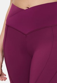 Des leggings taille haute en tissu violet profond avec une texture lisse et un design croisé au niveau de la taille, présentant des détails de couture contrastés.