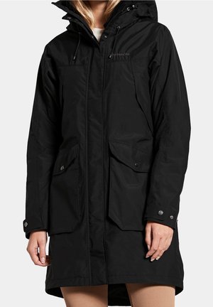 Femme portant un manteau d'hiver noir à capuche avec poches avant et poignets à boutons-pression, debout devant un fond clair uni.