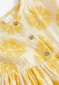 Vestido amarillo bordado con un patrón a rayas, que presenta botones de madera y un tejido recogido en la cintura, mostrando diseños florales.