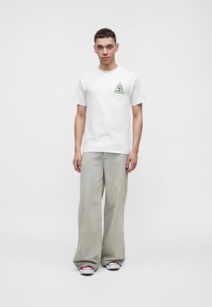 Junger Mann trägt ein weißes grafisches T-Shirt, lockere graue Jeans und rot-weiße Sneakers, steht vor einem schlichten hellen Hintergrund.