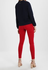 Felpa navy con scollo rotondo, abbinata a jeans skinny rossi. I sandali hanno cinturini alla caviglia e un tacco spesso. Texture del tessuto lisce visibili.