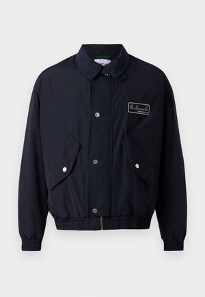 Navy blue jacket with a front zipper, snap buttons, two side pockets, and ribbed cuffs. Features a logo patch on the left chest area.