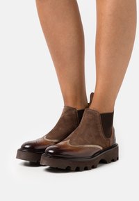 Stivali Chelsea in pelle e suede marrone con upper testurizzato, pannelli elastici laterali e una suola spessa e rigata con dettagli brogue decorativi.