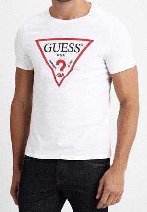 Mężczyzna w białej koszulce z czerwonym logo w kształcie trójkąta, na którym napisano "GUESS U.S.A. Washed Jeans" oraz w ciemnoniebieskich dżinsach.