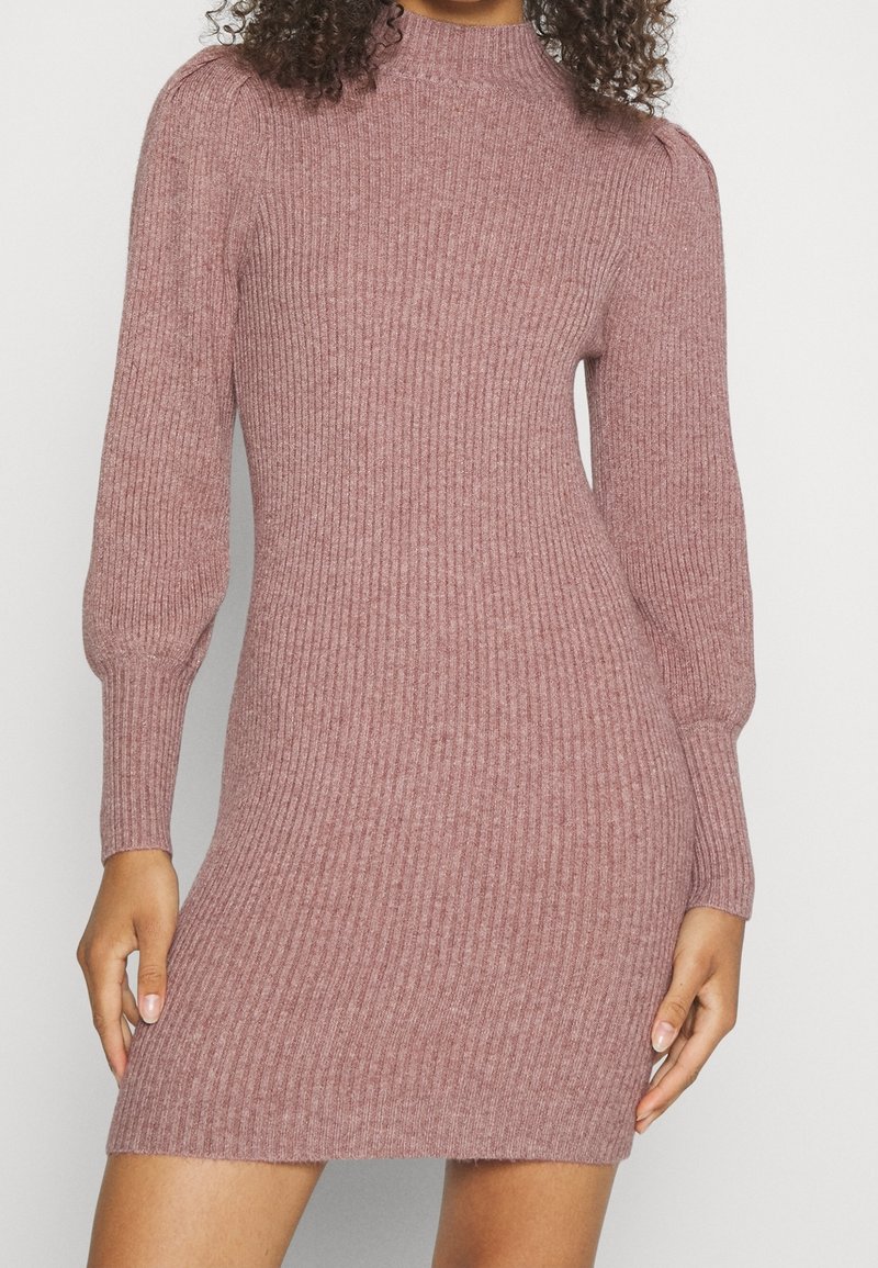 Femme portant une robe tricotée côtelée, longue jusqu'aux genoux, manches longues, couleur mauve, avec un col montant et des épaules légèrement bouffantes.