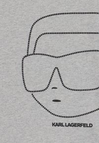 Pelēka t-kreklu ar melnu kontūru grafiku, kurā attēlotas saulesbrilles un seja, zem kuras melnās burtos ir rakstīts "KARL LAGERFELD".