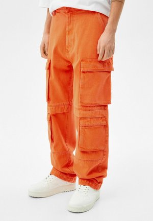 Persona con pantalones cargo sueltos de color naranja con múltiples bolsillos grandes y zapatillas blancas, de pie contra un fondo blanco.