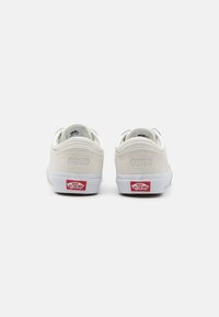 Zapatillas de ante blanco con un perfil bajo, que cuentan con una suela de goma blanca y una etiqueta de logo roja de Vans en el talón. Diseño minimalista y bordes sin costuras.