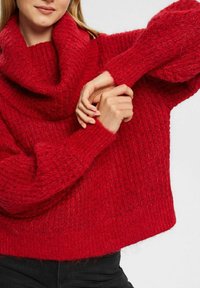 Roter gestrickter Pullover mit einem voluminösen Kragen, der mit strukturierten Mustern und gerippten Ärmeln versehen ist. Der Stoff wirkt warm und gemütlich.