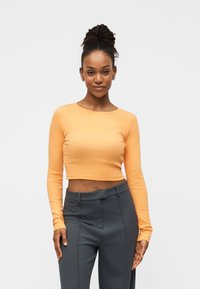 Μπλούζα με μακριά μανίκια και κομμένη γραμμή σε απαλό πορτοκαλί ύφασμα, συνδυασμένη με ψηλόμεσο, σκούρο γκρι παντελόνι με tailored εφαρμογή, που διαθέτει λείο ύφασμα και κοντινή γραμμή.