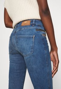 Jeans de denim azul con un ajuste ceñido, que cuentan con bolsillos traseros, tachuelas metálicas decorativas y un detalle de cremallera lateral. Parche de logo de cuero en la cinturilla.