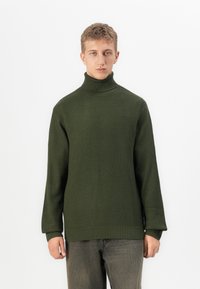 Junge Person in einem olivgrünen gestrickten Rollkragenpullover und dunkelgrauen Hosen, die vor einem schlichten weißen Hintergrund steht.