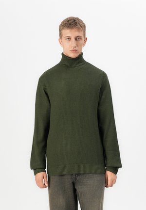 Jongere in een olijfgroene gebreide coltrui en donkergrijze broek, staand tegen een effen witte achtergrond.