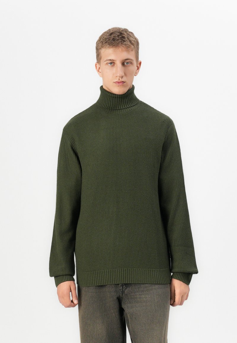 Junge Person in einem olivgrünen gestrickten Rollkragenpullover und dunkelgrauen Hosen, die vor einem schlichten weißen Hintergrund steht.