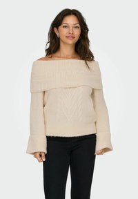 Sweter w kolorze kremowym z odkrytymi ramionami, o splocie w warkocze, z szerokimi rękawami i prążkowaną teksturą, połączony z czarnymi dżinsami.